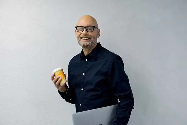 Mies hymyilee kannettava ja kahvimuki kädessä