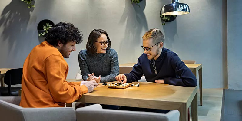 Kolme henkilöä pelaa lautapeliä pöydän ääressä