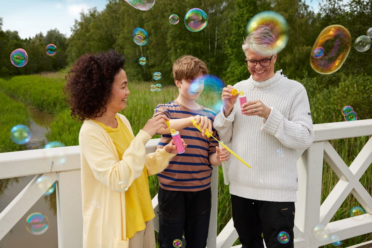 Kolme henkilöä puhaltaa saippuakuplia sillalla, taustalla metsää