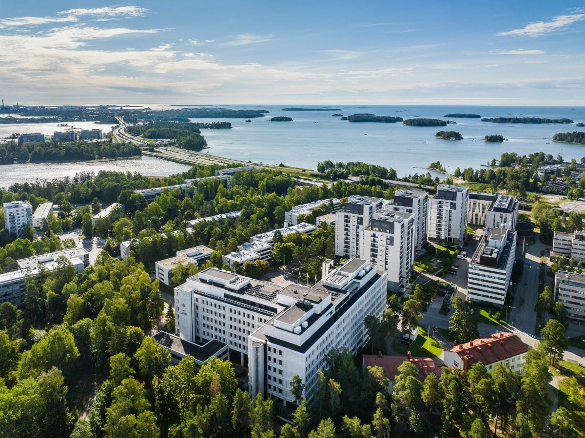 Ilmakuva toimitilarakennus Tapiolan Helmestä ja asuinrakennuksista vehreässä Espoossa. Taustalla kaunis merimaisema.