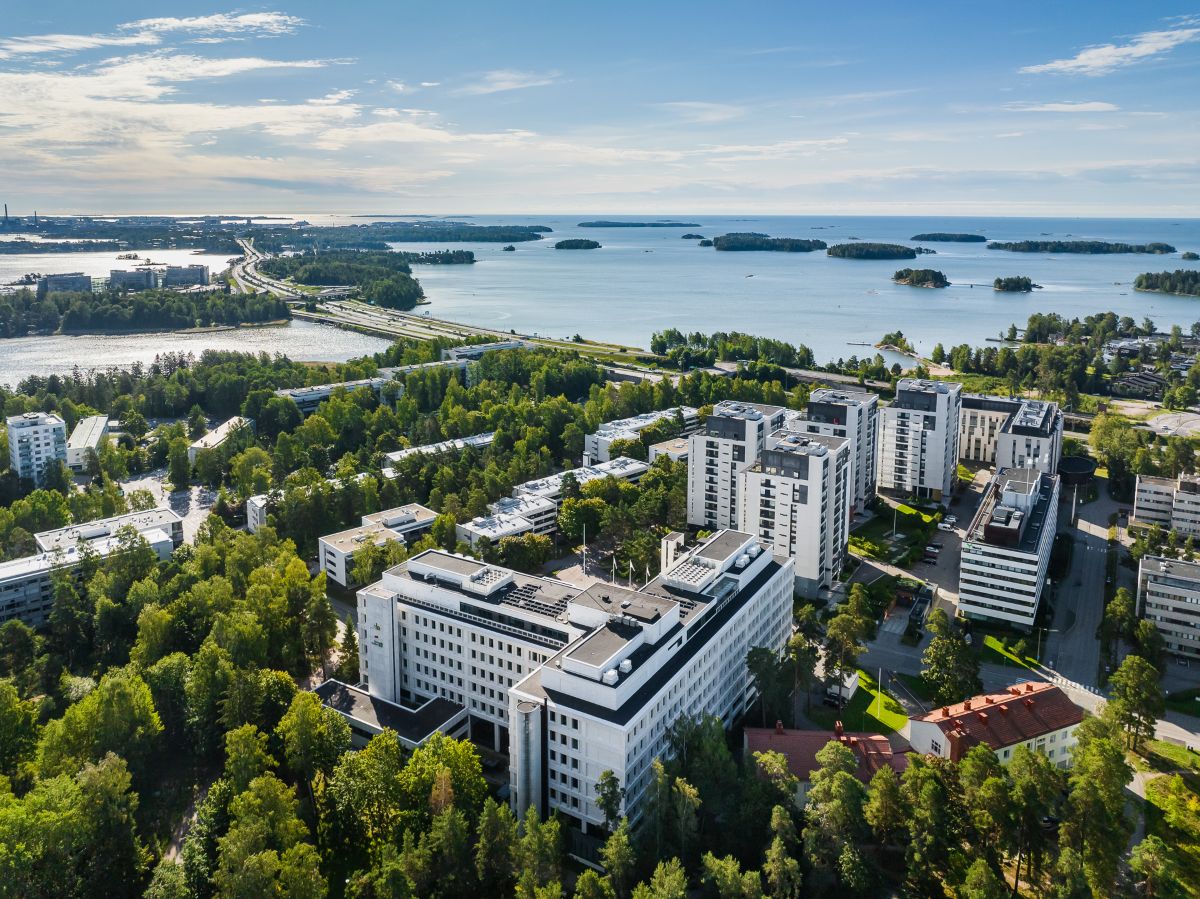 Ilmakuva toimitilarakennus Tapiolan Helmestä ja asuinrakennuksista vehreässä Espoossa. Taustalla kaunis merimaisema.