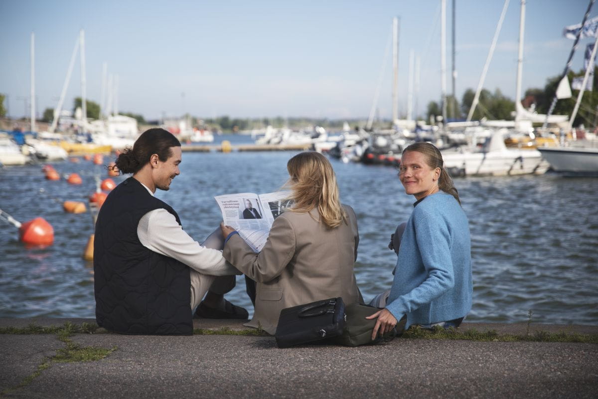 Kolme henkilöä selaa lehteä venesataman rannalla veneiden äärellä.